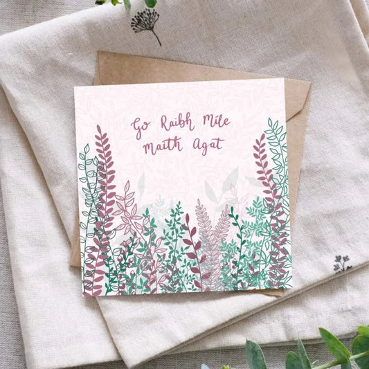 And Irish language thank you card that says "Go Raibh Míle Maith Agat" in Irish along the top of the card. Below the writing are bunches and branches of leaves, and buds in a mixture of dark pink and lots of green tones. Beneath the card is a recycled brown Kraft envelope and they are both sitting on a folded cloth surrounded by foliage for display.