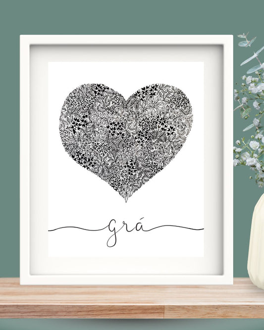 Framed Irish language art print. The black and white pen illustration of a heart is packed full of small intricate leaves. The botanical monochrome design is set against a white background and under the heart is the Irish word "Grá" which means love, in a flowing hand-written script. There is flowing line from each end of the word reaching out to the edges of the print. The print is set in a solid wood Irish made white frame and white mount. The frame is sitting on a wooden shelf against a deep green wall.