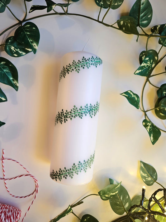 A white pillar candle with a hand-painted leafy design in green, placed on a surface with green leaves and red decorations, likely to be used as a centerpiece for Christmas.
