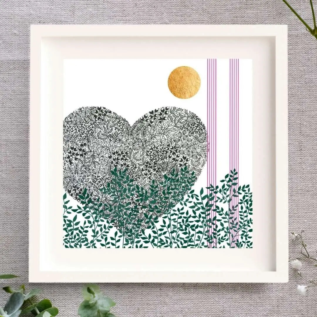 Image of a framed botanical art illustration. The frame is square, wooden, and white with a white mount. It is set on a linen cloth surrounded by baby's breath and eucalyptus.
The print is of a black pen illustration of a heart. Below the heart along the bottom of the image there are green leaves and branches. Up the right side of the print there are two strips of 5 pink lines, like music lines. At the top of the image there is a bright circle of hand-applied gold leaf.