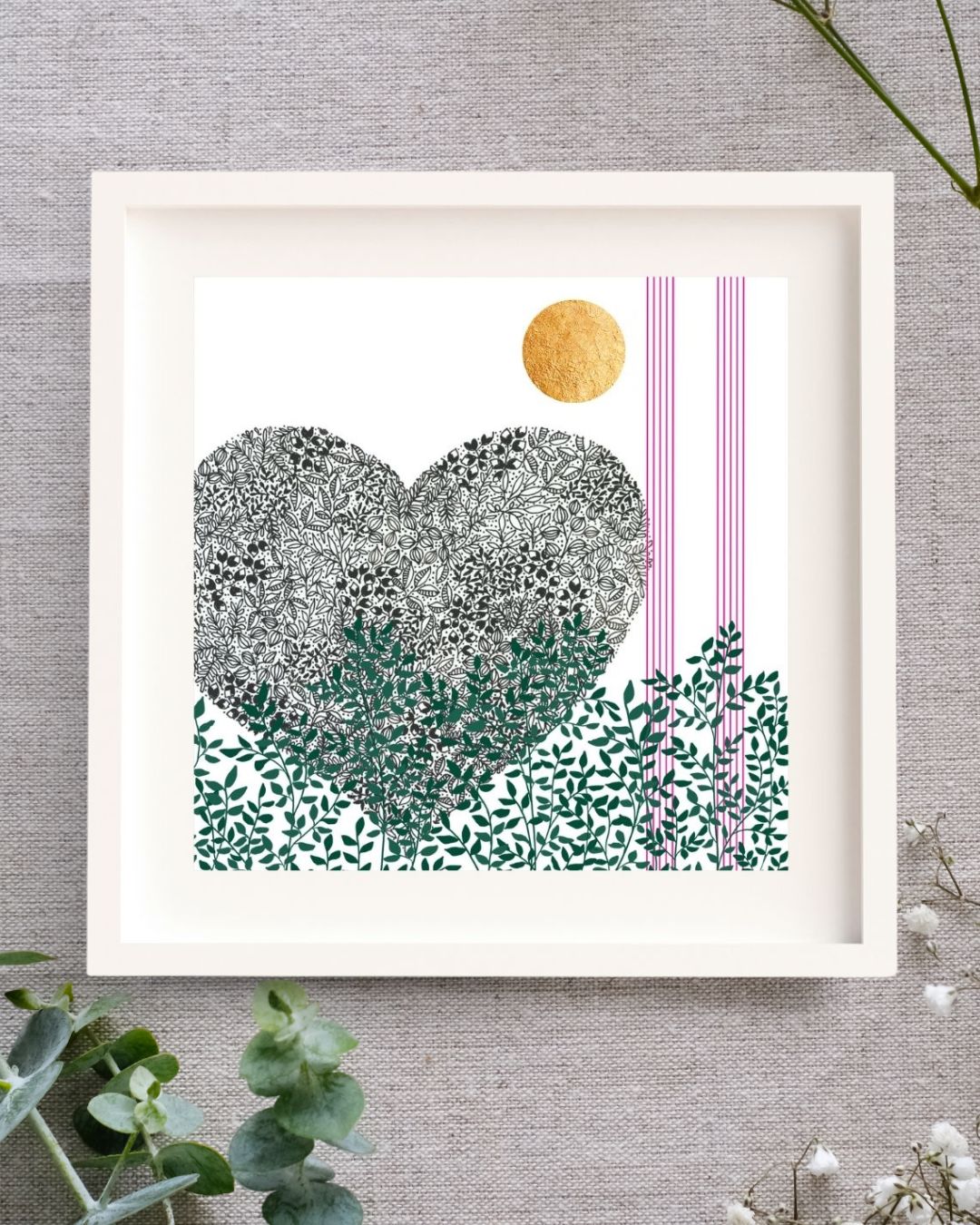 Image of a framed botanical art illustration. The frame is square, wooden, and white with a white mount. It is set on a linen cloth surrounded by baby's breath and eucalyptus.
The print is of a black pen illustration of a heart. Below the heart along the bottom of the image there are green leaves and branches. Up the right side of the print there are two strips of 5 pink lines, like music lines. At the top of the image there is a bright circle of hand-applied gold leaf.