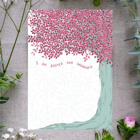 Botanical affirmation print with a tree on the right side of the design. The tree has a strong green trunk and is topped with lush leaves of various shades of pink. Underneath the leaves is the phrase "I am rooted and grounded"
The print is lying on a background of cloth and sprigs of greenery and flowers for decoration.