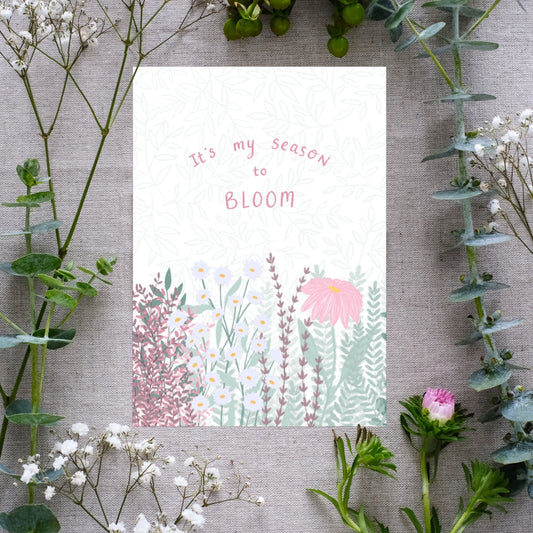 A botanical print titled 'It's my season to bloom', featuring illustrations of plants and flowers in various colors on a white background, surrounded by an array of real flowers and foliage.