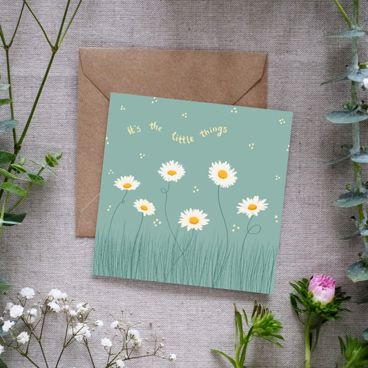 Image of a square greeting card with a muted green background, featuring a delicate illustration of white daisies with yellow centers growing in tall grass. Small scattered dots surround the flowers, adding a whimsical touch. Above the floral design, the phrase 'It's the little things' is written in soft yellow, curved text. The card is placed on a brown kraft envelope, resting on a textured linen surface, surrounded by sprigs of greenery and small white and pink flowers.