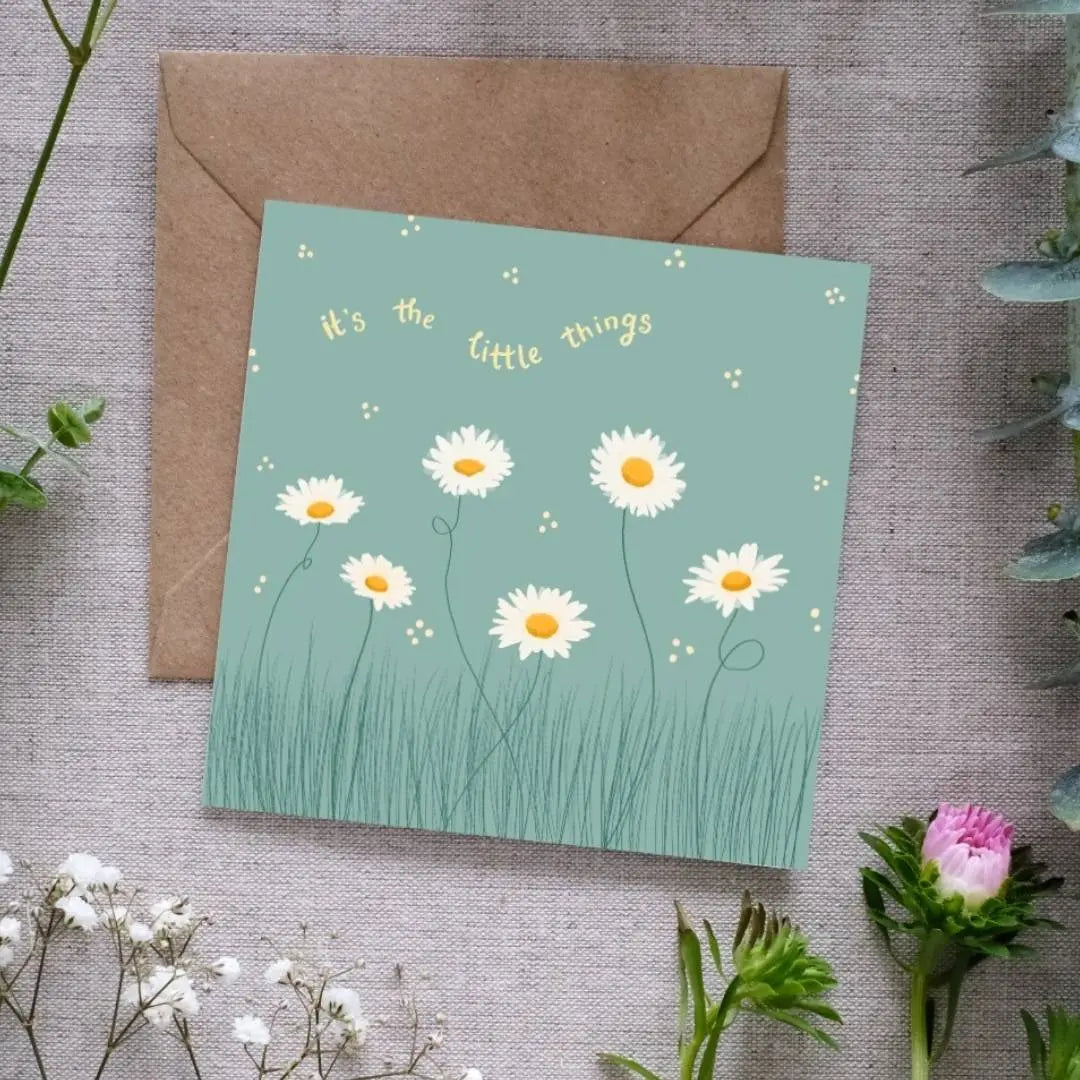 Image of a square greeting card with a muted green background, featuring a delicate illustration of white daisies with yellow centers growing in tall grass. Small scattered dots surround the flowers, adding a whimsical touch. Above the floral design, the phrase 'It's the little things' is written in soft yellow, curved text. The card is placed on a brown kraft envelope, resting on a textured linen surface, surrounded by sprigs of greenery and small white and pink flowers.