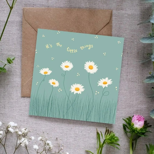 Image of a square greeting card with a muted green background, featuring a delicate illustration of white daisies with yellow centers growing in tall grass. Small scattered dots surround the flowers, adding a whimsical touch. Above the floral design, the phrase 'It's the little things' is written in soft yellow, curved text. The card is placed on a brown kraft envelope, resting on a textured linen surface, surrounded by sprigs of greenery and small white and pink flowers.