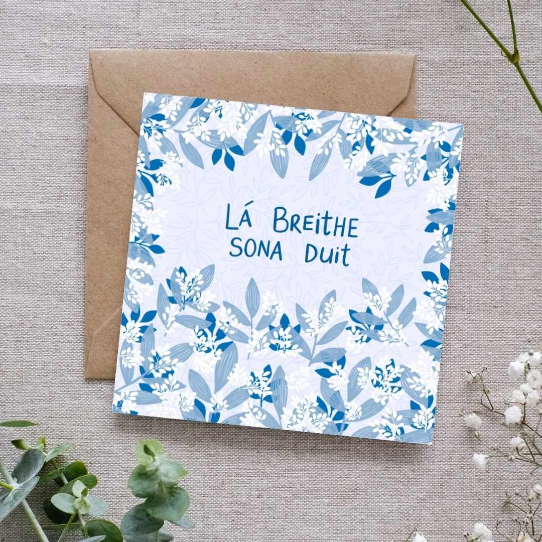 Irish language birthday card with a leafy botanical design in a mixture of blue tones intertwined with clusters of small white leaves. The text 'Lá Breithe Sona Duit' is hand written in the centre of the card. The birthday card is accompanied by a recycled brown Kraft envelope, displayed on a linen background with some green foliage at the bottom.