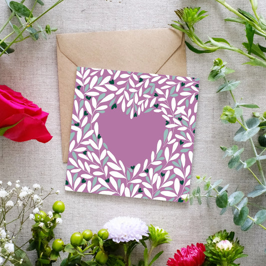 Square greeting card with a muted mauve background, featuring a heart-shaped cutout surrounded by an intricate leafy pattern in white, green, and dark accents. The delicate botanical design creates a romantic and elegant feel. The card is placed on a brown kraft envelope, resting on a textured fabric surface, surrounded by fresh flowers including red roses, white blossoms, and green foliage.