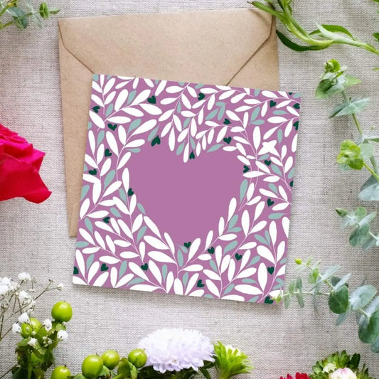 Square greeting card with a muted mauve background, featuring a heart-shaped cutout surrounded by an intricate leafy pattern in white, green, and dark accents. The delicate botanical design creates a romantic and elegant feel. The card is placed on a brown kraft envelope, resting on a textured fabric surface, surrounded by fresh flowers including red roses, white blossoms, and green foliage.
