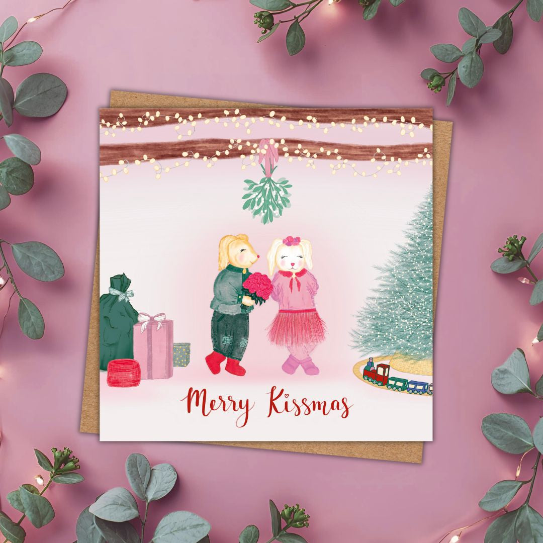A square Christmas card with a kraft envelope, styled on a dusty pink background with eucalyptus leaves and string lights. The card features a watercolor illustration of a sweet couple of woodland animals exchanging a pink bouquet under a mistletoe tree, with the text 'Merry Kissmas' at the bottom.
