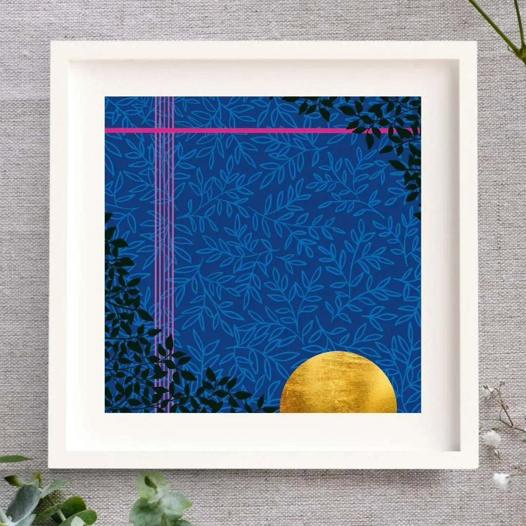 Midnight Magic is a framed art piece in a white mount and solid wood white frame. The illustration is moody and rich with a deep blue background, a subtle lighter blue leaf pattern layered on top, two striking pink lines, one at the top and one down the left side. There is a rich golden half sun/moon in gold leaf in the bottom right corner and there's some very dark green leaves in a triangle in the bottom left corner and top right corner. The frame is laid on a cloth with some foliage surrounding it.