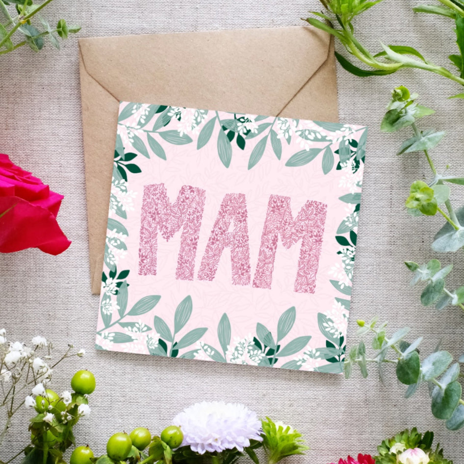 Square greeting card with the word 'MAM' in large pink floral-patterned letters on a soft pink background. The card is decorated with a border of green leaves and small white flowers. A brown kraft envelope is partially visible behind the card. The card is lying on linen cloth with greenery and flowers scattered around it.