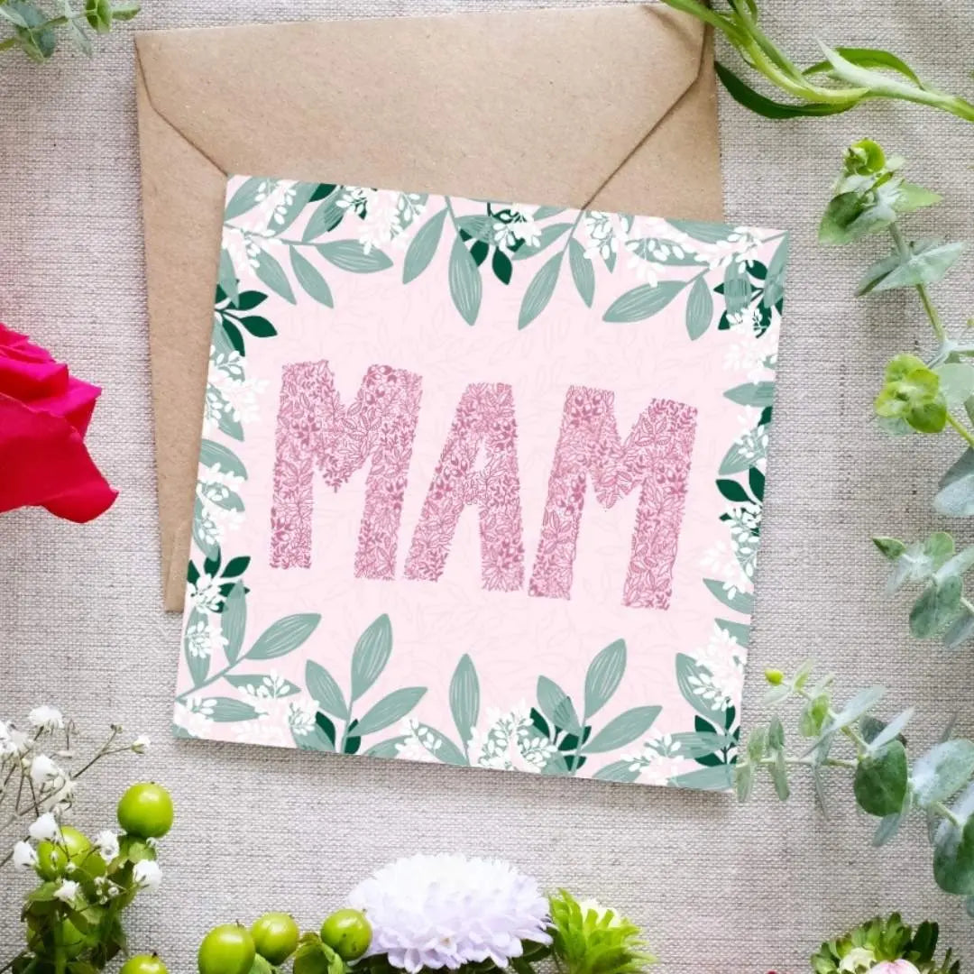 Square greeting card with the word 'MAM' in large pink floral-patterned letters on a soft pink background. The card is decorated with a border of green leaves and small white flowers. A brown kraft envelope is partially visible behind the card. The card is lying on linen cloth with greenery and flowers scattered around it.