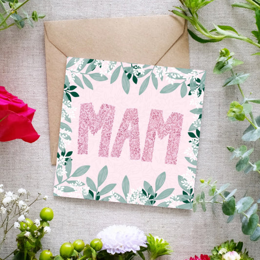 Square greeting card with the word 'MAM' in large pink floral-patterned letters on a soft pink background. The card is decorated with a border of green leaves and small white flowers. A brown kraft envelope is partially visible behind the card. The card is lying on linen cloth with greenery and flowers scattered around it.