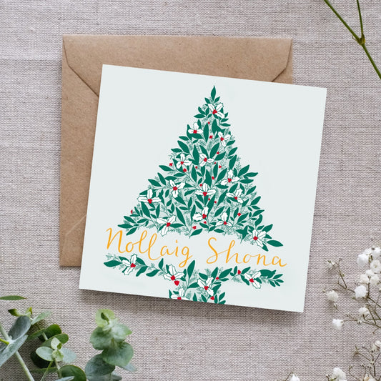 A Christmas tree greeting card with Irish-language text 'Nollaig Shona' and an illustration of a Christmas tree. The tree is made of green leaves, red berries, smaller intricate details white holly leaves. The background of the card is a very subtle soft green. The card is placed on a brown envelope and some decorative foliage in the background.