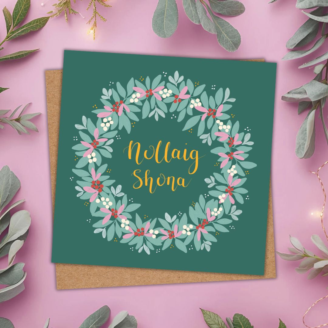 A square greeting card with a floral wreath design and the text 'Nollaig Shona' in gold script, placed on a brown kraft envelope against a pink background with decorative greenery.