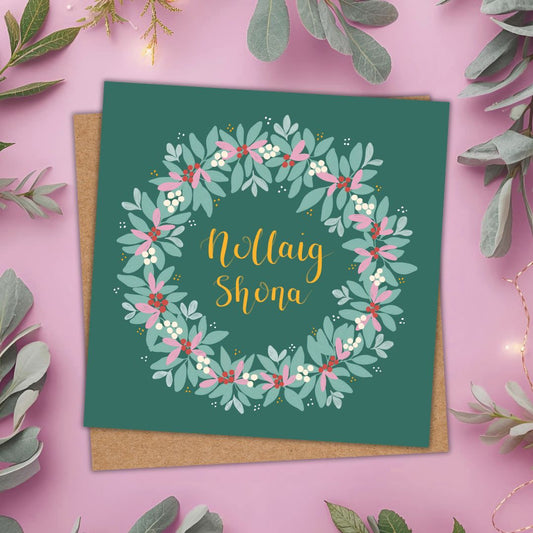 A square greeting card with a floral wreath design and the text 'Nollaig Shona' in gold script, placed on a brown kraft envelope against a pink background with decorative greenery.