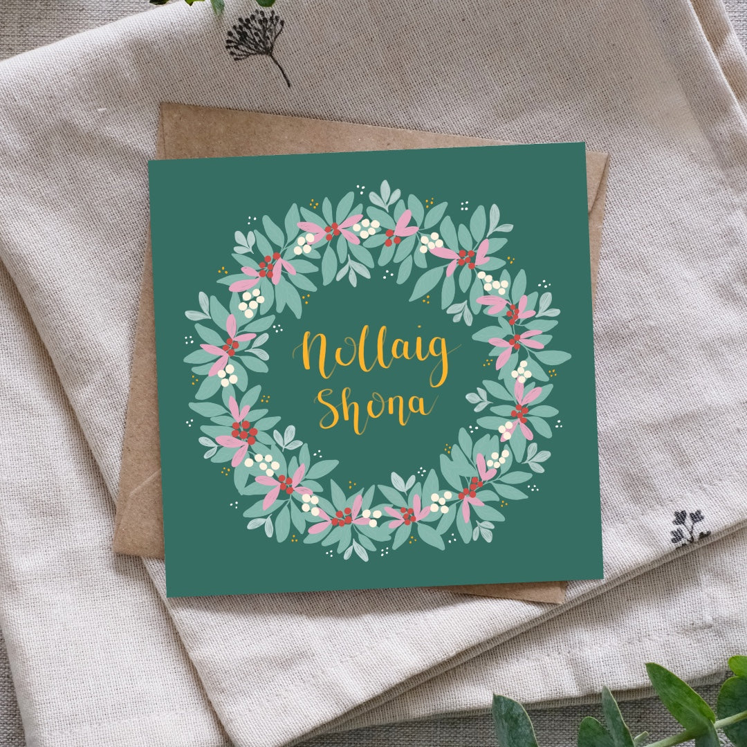 A square greeting card with a teal background and a floral wreath design, placed on top of a kraft envelope and beige fabric. The card reads 'Nollaig Shona' in gold script.