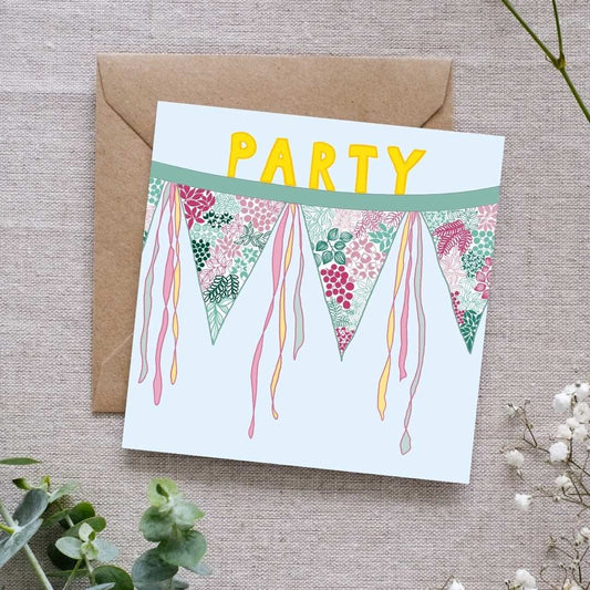 Square greeting card card with a pastel blue background, featuring colorful floral bunting flags with pink and green leafy patterns, accented by flowing ribbon tails. The word 'PARTY' is displayed in bold yellow letters at the top. The card is placed on a brown kraft envelope, set on a textured linen fabric surface, surrounded by delicate greenery and white flowers.