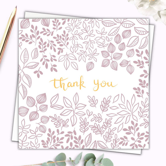 Pretty botanical Thank you card with dusty pink floral details and leaves and petals surrounding the words "Thank you" which is written in gold hand writing. The background of the botanical themed card is white so the details are clear and simple. The card is set on top of a plain white envelope.