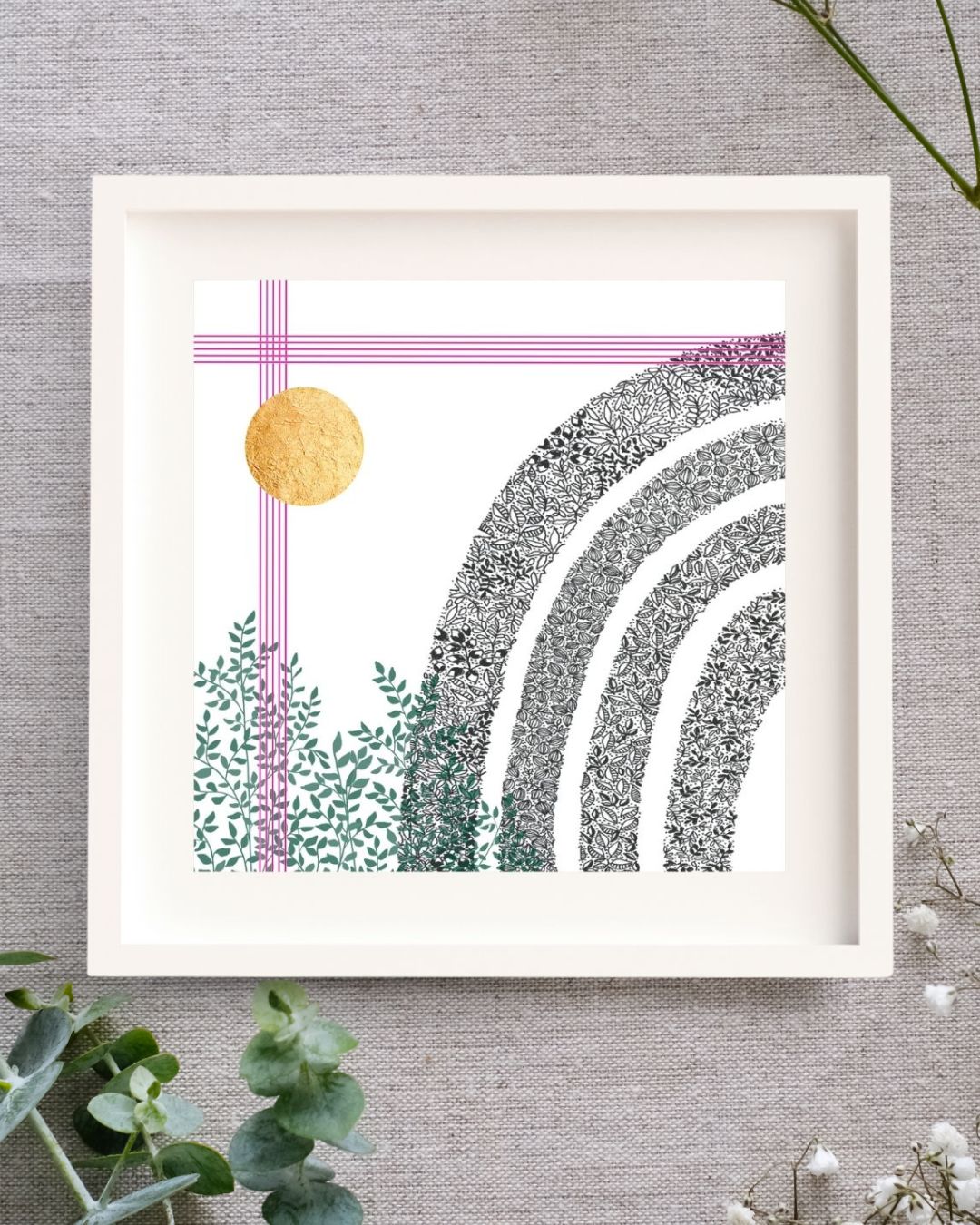 Image shows a framed art print lying on a linen cloth with sprinkles of baby's breath and eucalyptus around it. The frame is square and white. There is a white mount. The print is of a black and white pen illustration of a rainbow made of leaves. There's a cluster of green leaves in the bottom left corner. Straight up from there, there are five bright pink lines close together like music lines. The same 5 pink lines go across the top of the image, and lastly, there is a circle of hand-applied gold leaf.