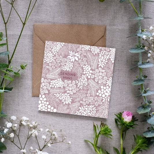 A square greeting card with a muted mauve background and intricate white floral and leafy patterns. The words 'SENDING HUGS' are centered in small, hand-written font. The card is placed on a textured linen surface, partially resting on a brown kraft envelope. Surrounding the card are sprigs of eucalyptus, baby’s breath, and other greenery, along with a small pink flower, for presentation.