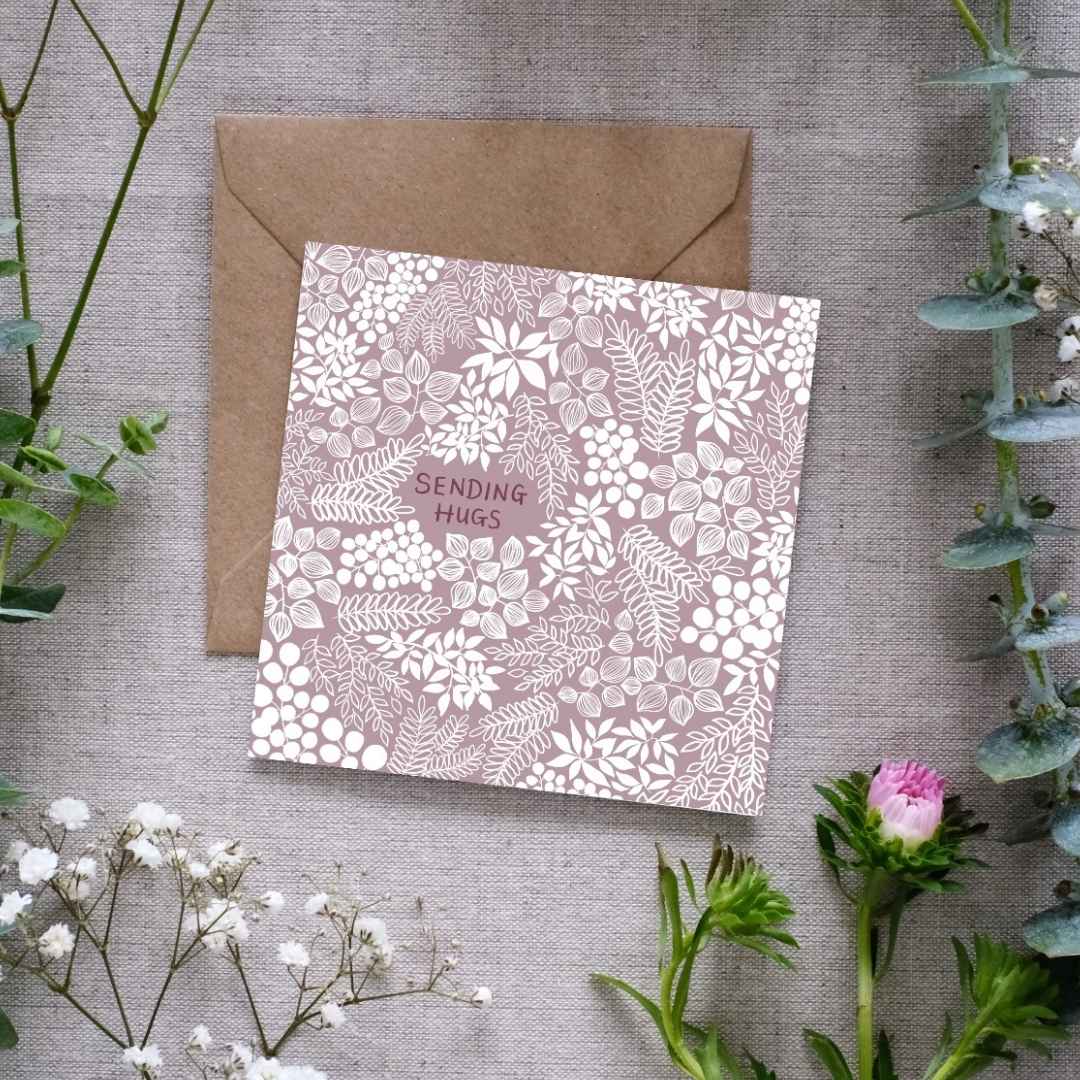 A square greeting card with a muted mauve background and intricate white floral and leafy patterns. The words 'SENDING HUGS' are centered in small, hand-written font. The card is placed on a textured linen surface, partially resting on a brown kraft envelope. Surrounding the card are sprigs of eucalyptus, baby’s breath, and other greenery, along with a small pink flower, for presentation.