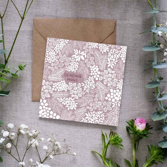 A square greeting card with a muted mauve background and intricate white floral and leafy patterns. The words 'SENDING HUGS' are centered in small, hand-written font. The card is placed on a textured linen surface, partially resting on a brown kraft envelope. Surrounding the card are sprigs of eucalyptus, baby’s breath, and other greenery, along with a small pink flower, for presentation.