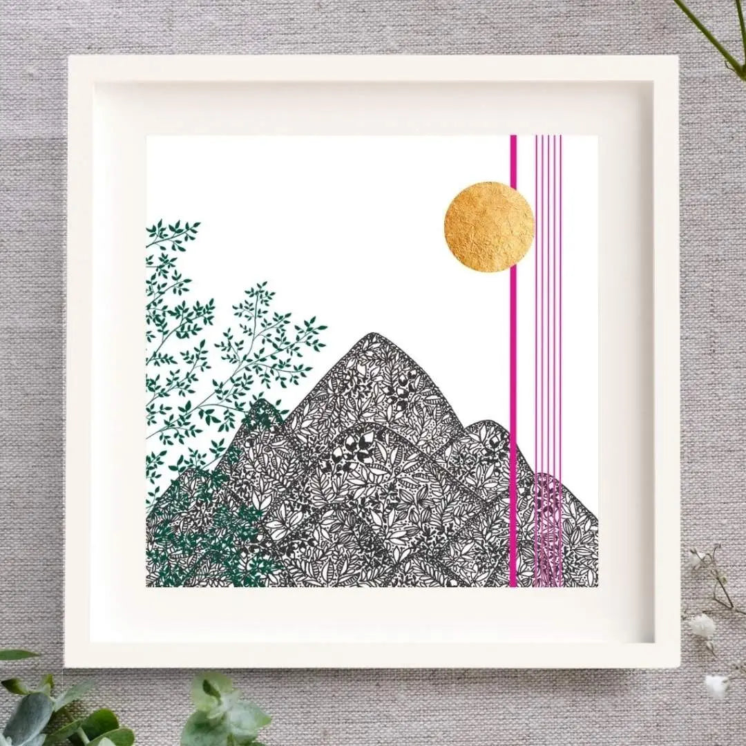 Image of a framed art print featuring a botanical black pen illustration of mountains made up of intricate leaf patterns. Green leafy branches climb up the left side, while a gold leaf sun and thin pink vertical lines accent the right side. The artwork is set against a white background, a white mount, and framed in a simple white wooden frame, placed on a neutral fabric surface with eucalyptus leaves and baby's breath in the foreground.