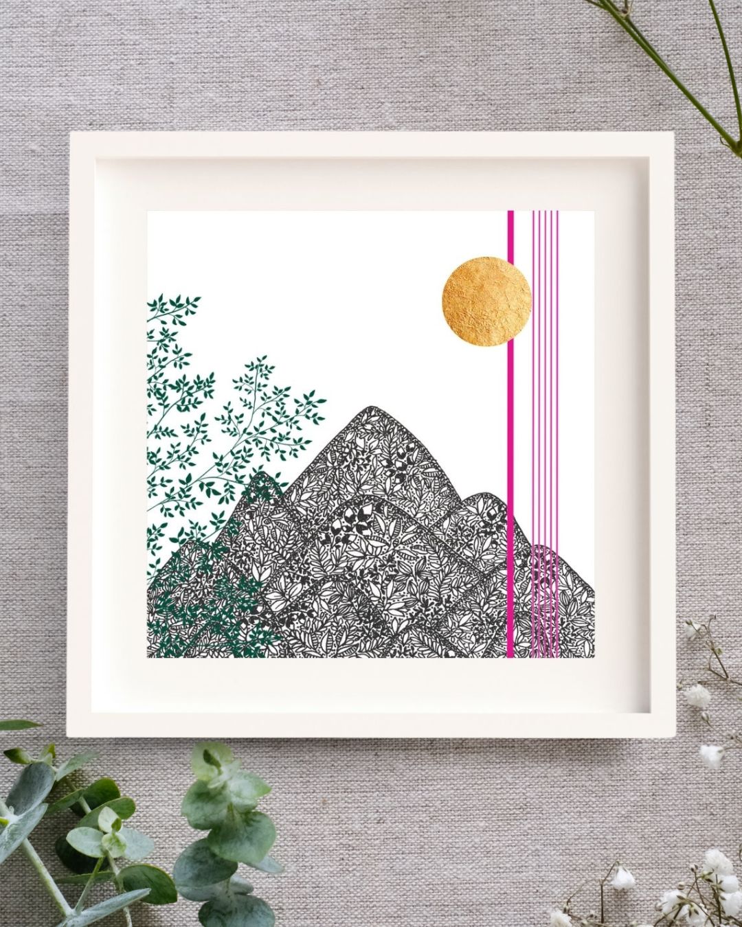 Image of a framed art print featuring a botanical black pen illustration of mountains made up of intricate leaf patterns. Green leafy branches climb up the left side, while a gold leaf sun and thin pink vertical lines accent the right side. The artwork is set against a white background, a white mount, and framed in a simple white wooden frame, placed on a neutral fabric surface with eucalyptus leaves and baby's breath in the foreground.