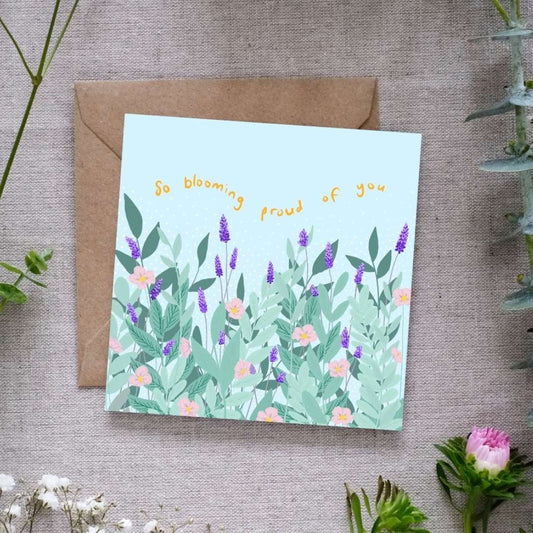 Image of a square greeting card with a pastel blue background, featuring an illustrated garden scene with lush green leaves, purple lavender stalks, and delicate pink flowers. Above the floral design, the phrase 'So blooming proud of you' is written in playful, curved yellow text. The card is placed on a brown kraft envelope, resting on a textured fabric surface, surrounded by sprigs of greenery and small pink and white flowers.