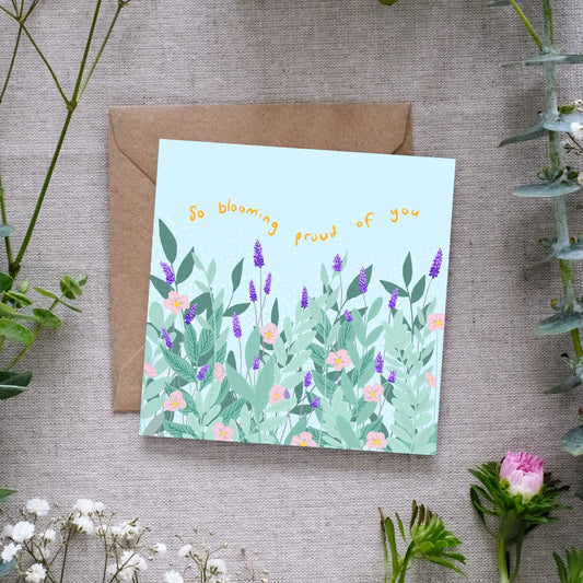 Image of a square greeting card with a pastel blue background, featuring an illustrated garden scene with lush green leaves, purple lavender stalks, and delicate pink flowers. Above the floral design, the phrase 'So blooming proud of you' is written in playful, curved yellow text. The card is placed on a brown kraft envelope, resting on a textured fabric surface, surrounded by sprigs of greenery and small pink and white flowers.