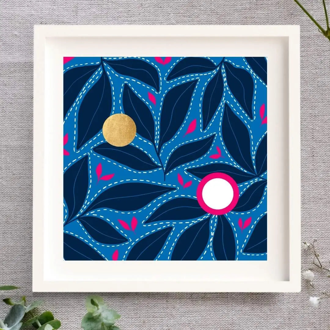 Framed Irish artwork, Solas na Mara, showing dark blue leaf patterns floating on a light blue background. There are bring pink leaf accents in between the dark leaves. Above these are two circles. One is made from gold leaf so there's a shimmer to it, and the other is bright white, with a strong bight pink surround. The square print is framed in a solid wood white frame, laid out on a natural cloth, and there are sprinklings of leaves and buds nearby.