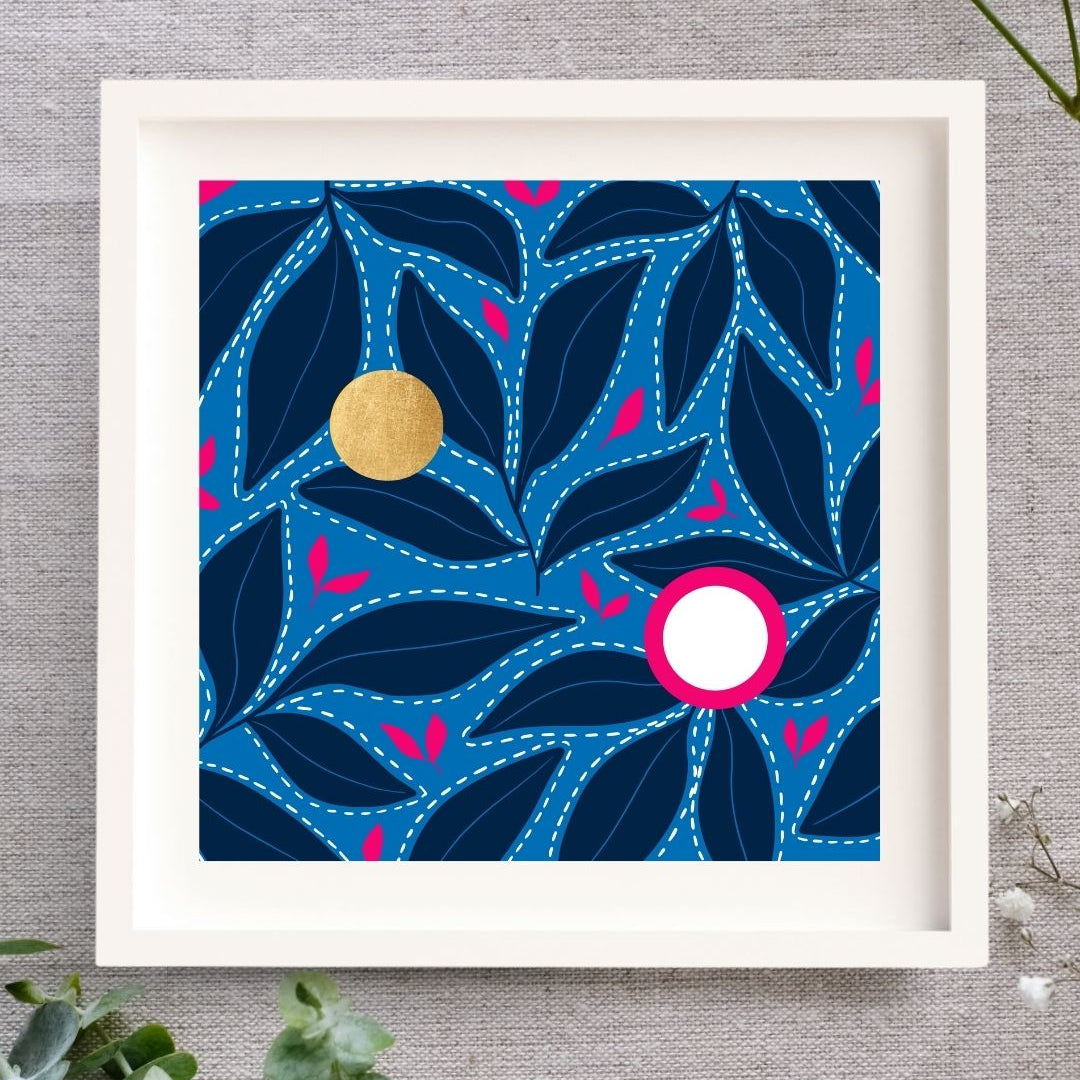 Framed Irish artwork, Solas na Mara, showing dark blue leaf patterns floating on a light blue background. There are bring pink leaf accents in between the dark leaves. Above these are two circles. One is made from gold leaf so there's a shimmer to it, and the other is bright white, with a strong bight pink surround. The square print is framed in a solid wood white frame, laid out on a natural cloth, and there are sprinklings of leaves and buds nearby.