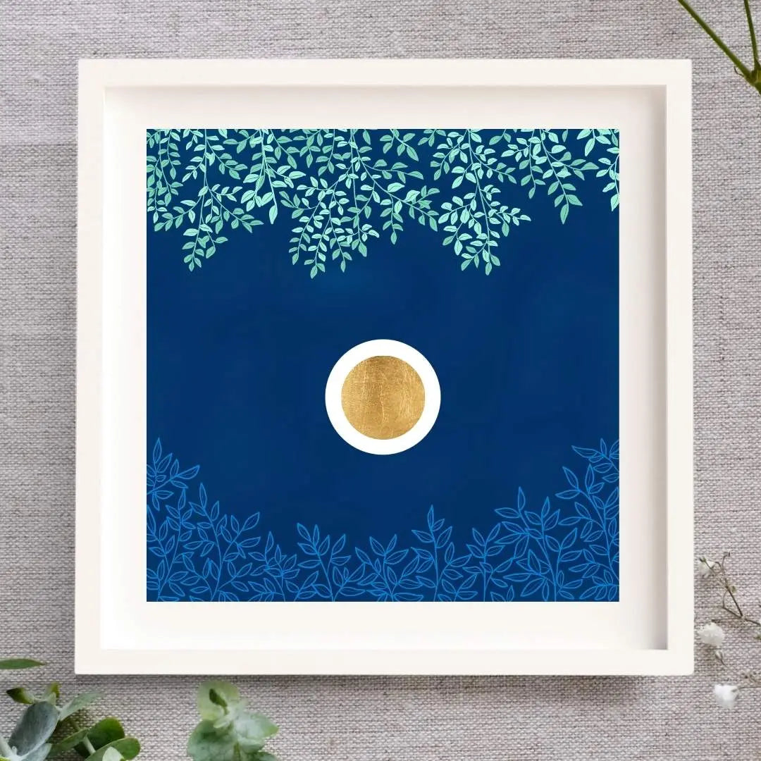 Framed artwork with a gold leaf circle in a white orb set in a dark blue textured sky. Above there are green branches, and below there are light blue. The Irish nature inspired art print is framed in a white solid wood frame, lying on a natural cloth surrounded by leaves and buds.