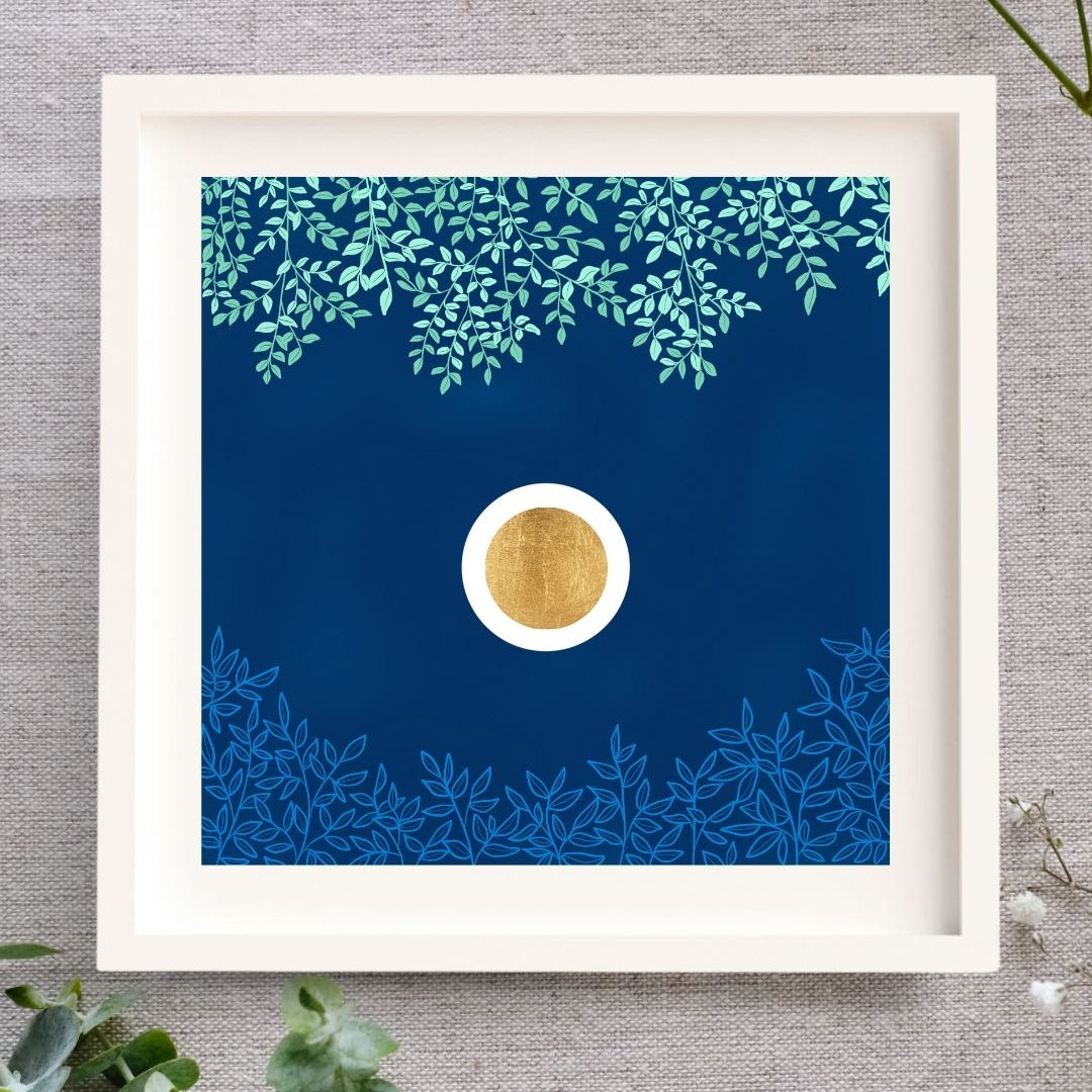 Framed artwork with a gold leaf circle in a white orb set in a dark blue textured sky. Above there are green branches, and below there are light blue. The Irish nature inspired art print is framed in a white solid wood frame, lying on a natural cloth surrounded by leaves and buds.