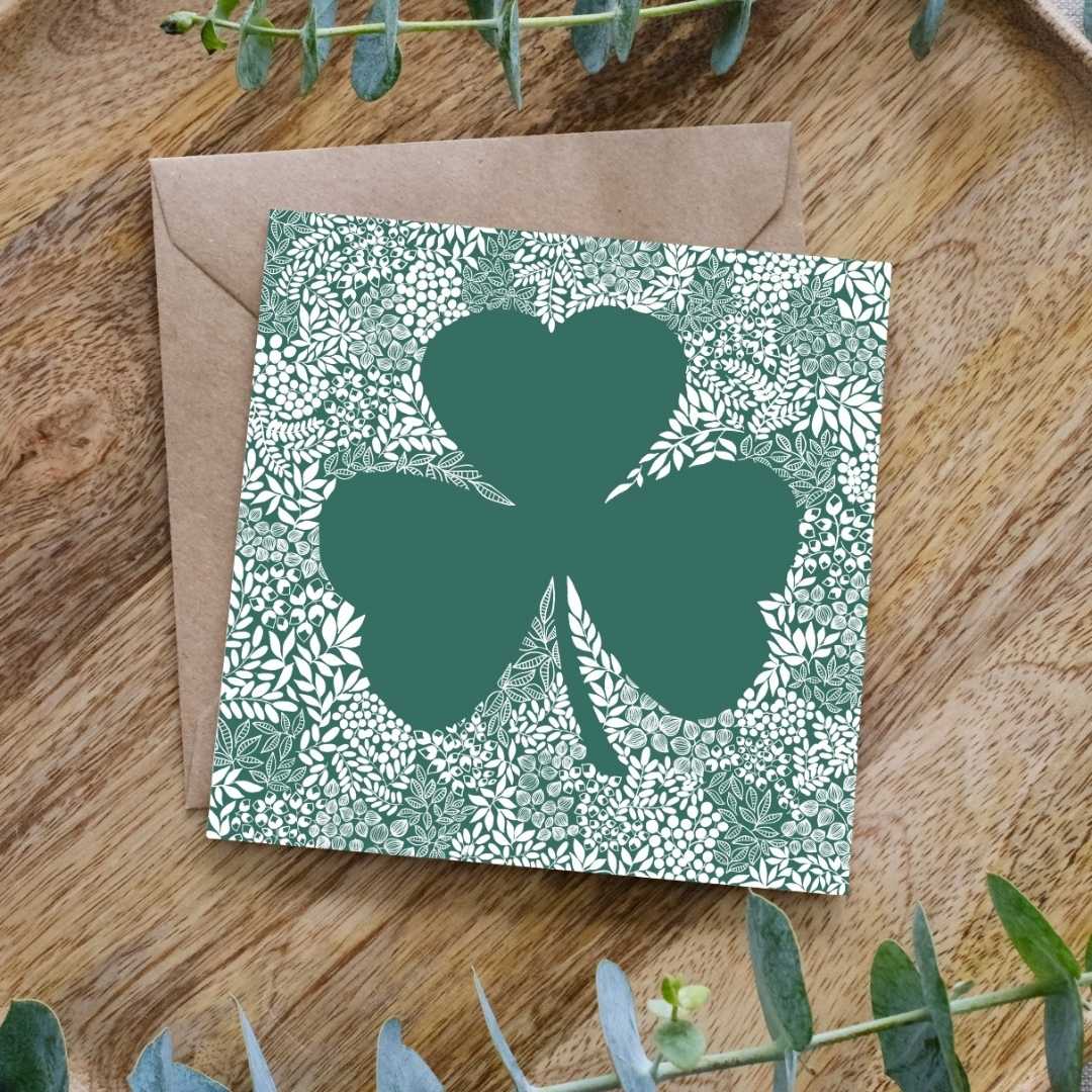 Image shows a square greeting card featuring a large green shamrock silhouette surrounded by an intricate white floral and leaf pattern. The detailed botanical design contrasts against the deep green background, creating a striking visual effect. The card is placed on a brown kraft envelope, resting on a wooden surface, with sprigs of eucalyptus leaves framing the scene.