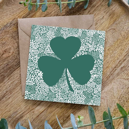 Image shows a square greeting card featuring a large green shamrock silhouette surrounded by an intricate white floral and leaf pattern. The detailed botanical design contrasts against the deep green background, creating a striking visual effect. The card is placed on a brown kraft envelope, resting on a wooden surface, with sprigs of eucalyptus leaves framing the scene.