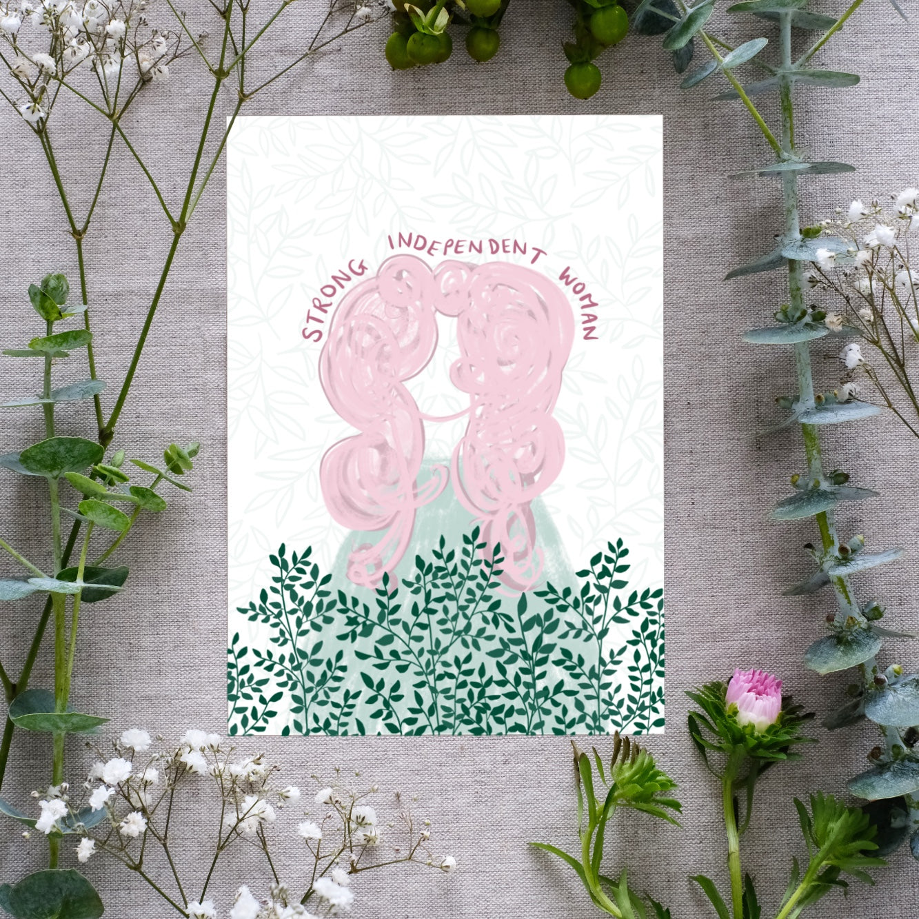 A floral affirmation print with the text 'Strong Independent Woman' written along the top of a feminine figure with large pink hair. Beneath the girl are sprigs of dark green greenery on a white background. The affirmation print is surrounded by real flowers for presentation.