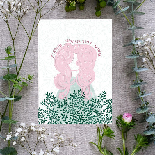A floral affirmation print with the text 'Strong Independent Woman' written along the top of a feminine figure with large pink hair. Beneath the girl are sprigs of dark green greenery on a white background. The affirmation print is surrounded by real flowers for presentation.