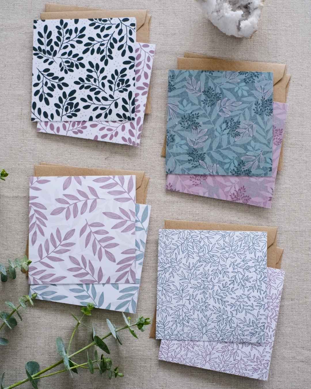 A collection of 8 square botanical-themed note cards, each featuring intricate leafy patterns in green and pink color schemes. The cards are stacked with matching brown kraft envelopes and laid out on a neutral natural fabric surface. Adding more natural tones to the image are sprigs of eucalyptus and greenery and a white crystal.