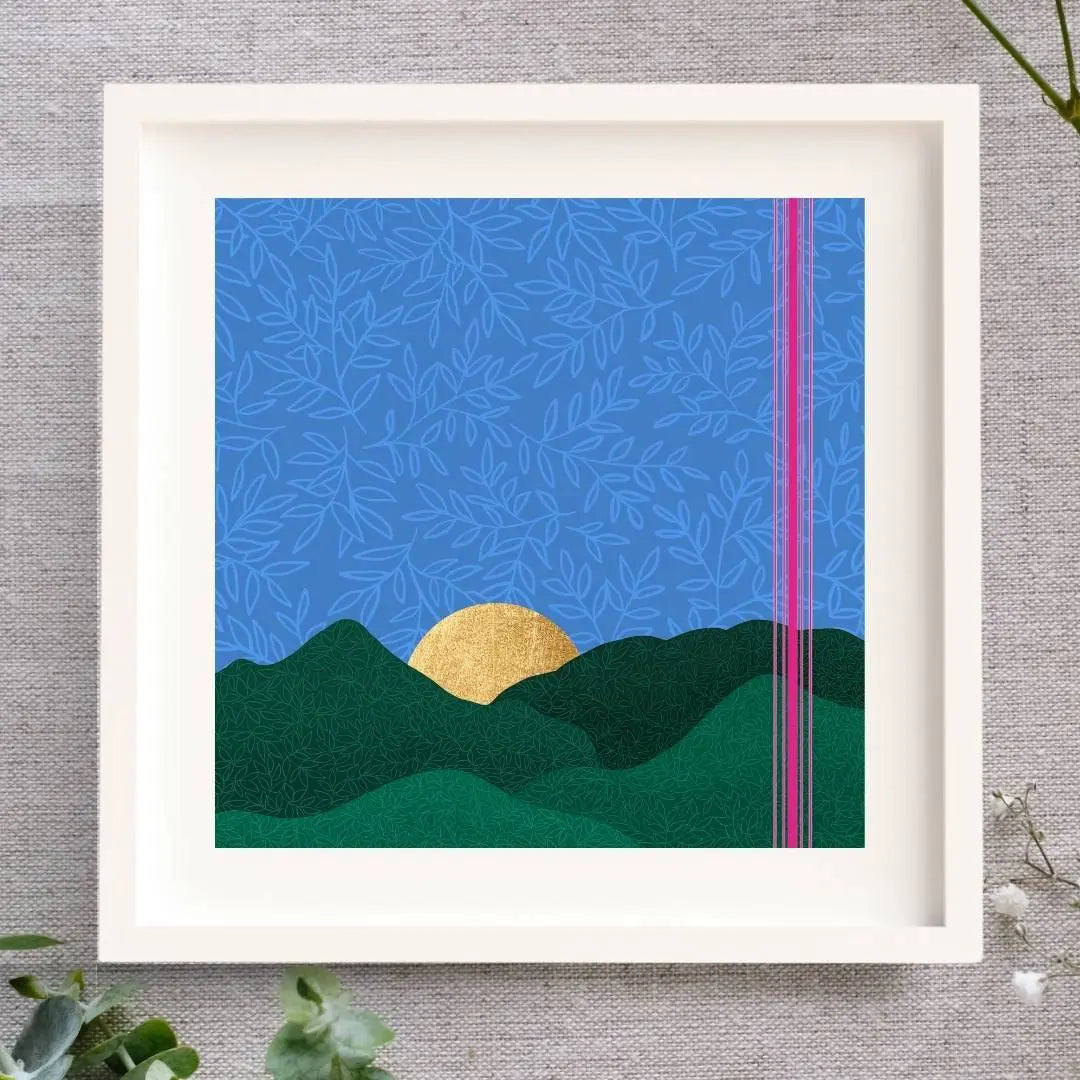 Framed artwork featuring an evening blue sky. There's a rich blue background with a leaf pattern layered over it. There are rolling layered hills and the sun is setting behind them. The sun is made from gold leaf. To the right there are 5 bright pink lines. The print is set in a mount and white frame, laid on a natural cloth.