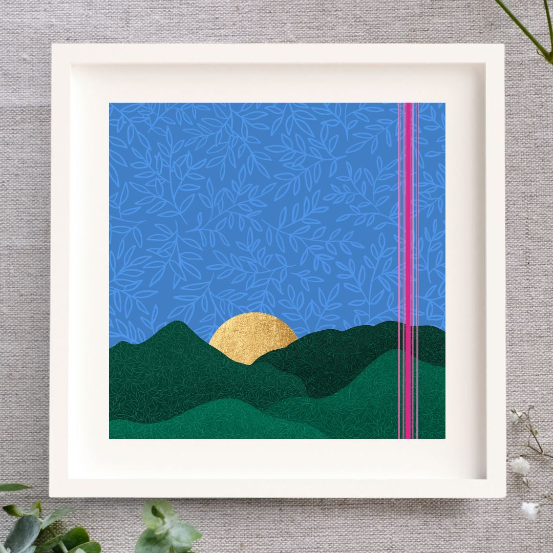 Framed artwork featuring an evening blue sky. There's a rich blue background with a softer lighter blue leaf pattern layered over it to give a velvety feel. There are rolling layered hills in green and the sun is setting behind them. The sun is made from gold leaf so there's a soft glow glimmer to it. To the right there is a cluster of bright pink lines going from the top of the print to the bottom. The illustration is set in a white mount and a white frame. It's laid on a natural cloth.