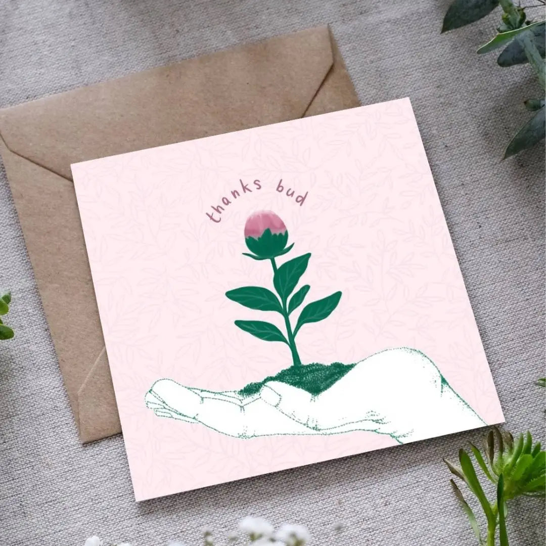 A greeting card with the outline of a hand facing upwards with a little green seedling with a pink bud at the top, sitting in the palm of the hand. The text 'Thanks bud' is written in dusty pink hand writing around the bud of the seedling. The card is accompanied by a brown envelope, displayed on a fabric surface with foliage around.