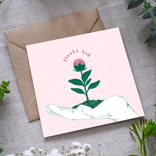 A greeting card with the outline of a hand facing upwards with a little green seedling with a pink bud at the top, sitting in the palm of the hand. The text 'Thanks bud' is written in dusty pink hand writing around the bud of the seedling. The card is accompanied by a brown envelope, displayed on a fabric surface with foliage around.