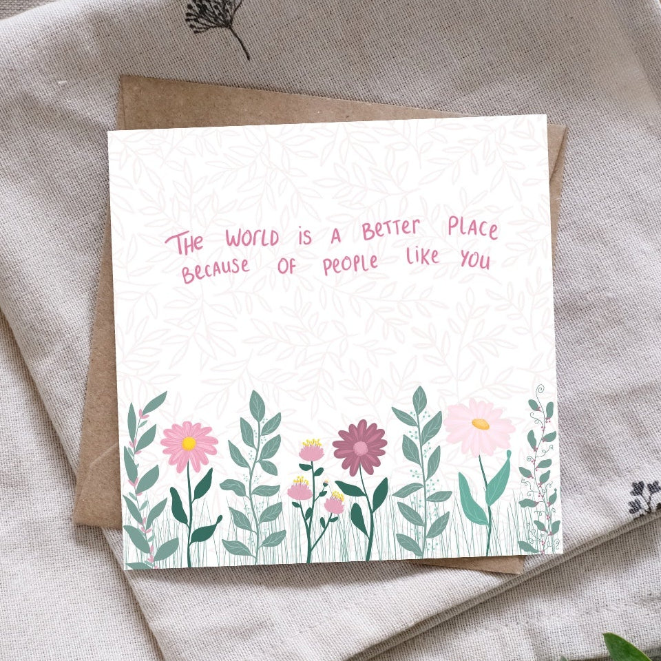 A square greeting card with a floral border and the quote 'THE WORLD IS A BETTER PLACE BECAUSE OF PEOPLE LIKE YOU' in pink text, placed on top of folded beige fabric with a kraft envelope. Green eucalyptus stems are arranged around it on a textured gray surface.