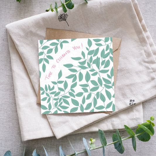 A square greeting card with a botanical leaf pattern in soft green tones on a white background. The card features our handwritten words "Time to Celebrate You!" in pink. It is placed on top of a brown kraft paper envelope, which rests on a folded linen cloth. Eucalyptus sprigs surround the arrangement, adding a natural touch.