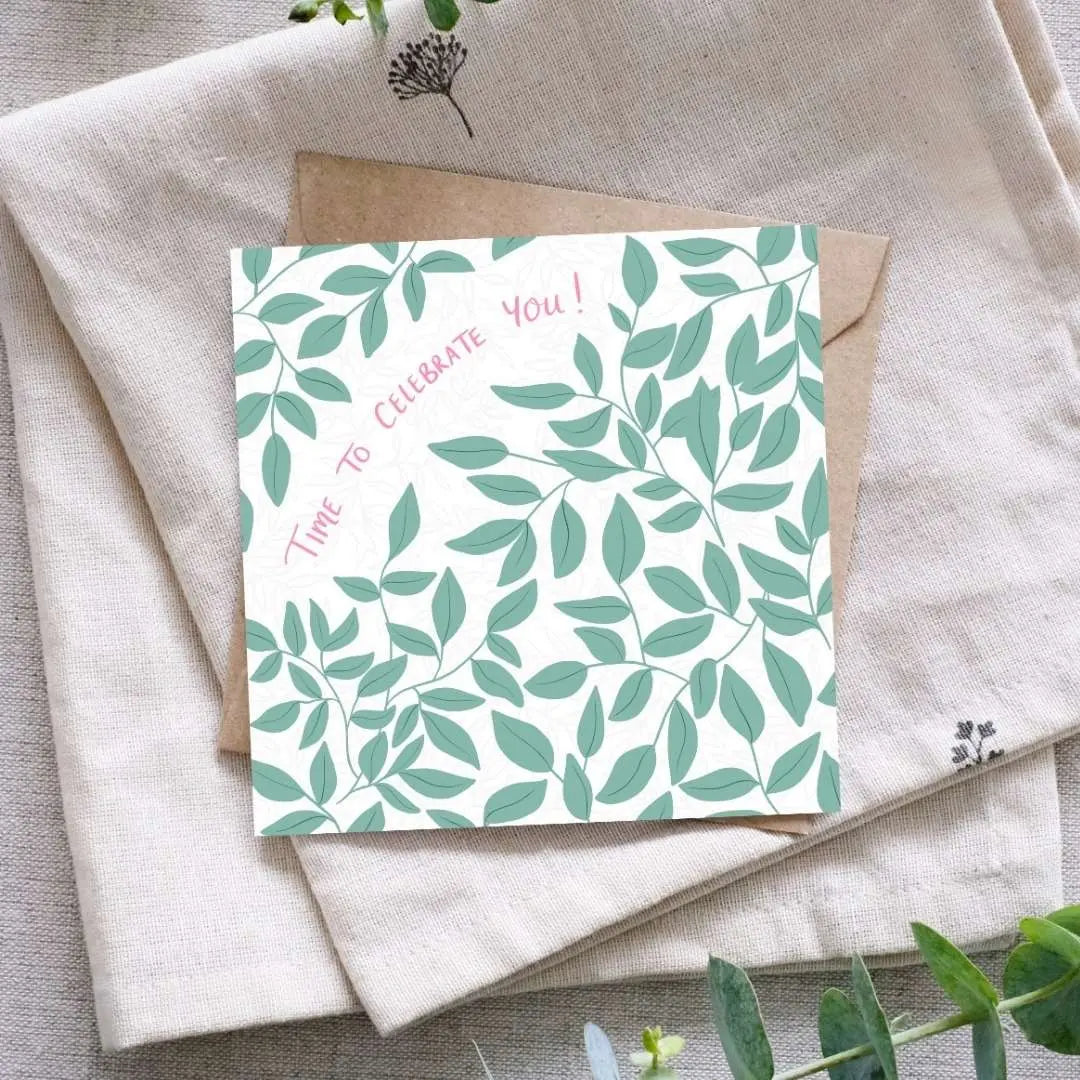 A square greeting card with a botanical leaf pattern in soft green tones on a white background. The card features our handwritten words "Time to Celebrate You!" in pink. It is placed on top of a brown kraft paper envelope, which rests on a folded linen cloth. Eucalyptus sprigs surround the arrangement, adding a natural touch.