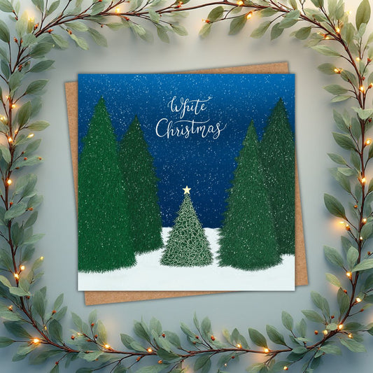 Christmas card with large green trees surrounding one small little tree that's lit up and has a gold star on top. The scene is set in a forest with a deep blue sky and snow falling. Above, 'White Christmas' is written in Calligraphy hand writing.