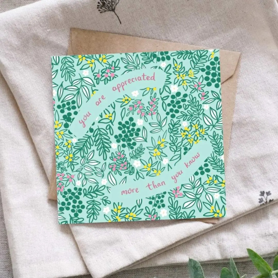 Image shows a pretty thank you card which says You Are Appreciated More Than You Know written in deep pink hand writing. The background of the card is a soft light green with dark green leaves, buds and floral details. There are scatterings of yellow and pink bud clusters as well as pretty daisy heads in between gaps in the leaves. The card is laid on a brown recycled Kraft envelope. Underneath that is a linen cloth for display.